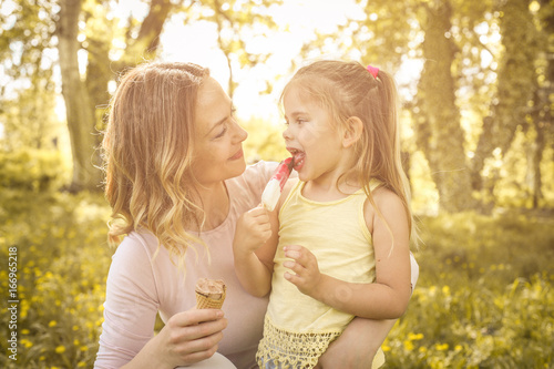 Mother and daughter sitting on the grass and eating ice cream. Happy mother with her daughter in the meadow.
