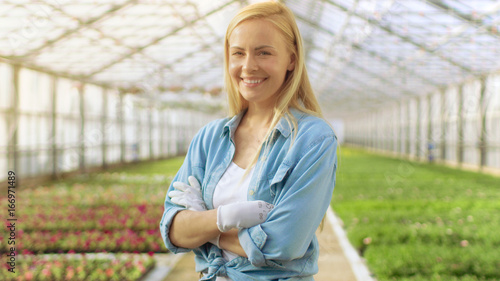 On a Sunny Day Beautiful Blonde Gardener Stands Smiling in a Greenhouse Full of Colorful Flowers.