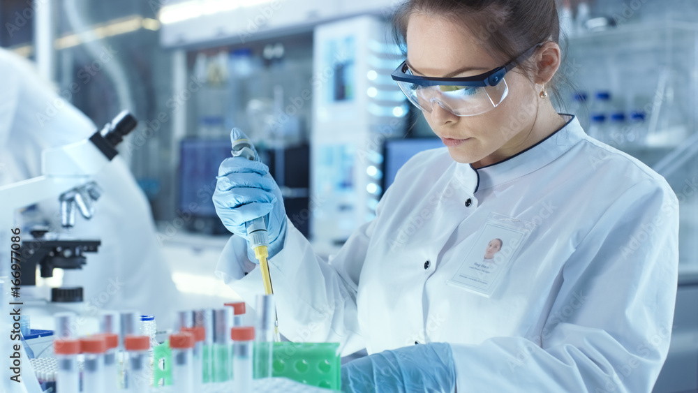 Female Research Scientist Uses Micropipette Filling Test Tubes in a Big Modern Laboratory. In the Background Scientists are Working. Female Research Scientist Uses Micropipette Filling Test Tubes in a Big Modern Laboratory. In the Background Scientists are Working.