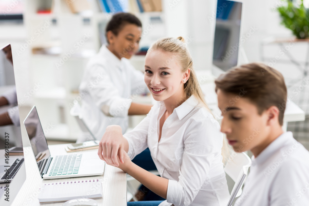 selective focus of smiling teenage girl looking at camera while studying with classmates in class selective focus of smiling teenage girl looking at camera while studying with classmates in class