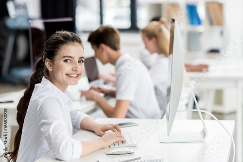 side view of smiling teenage girl looking at camera while typing on computer keyboard