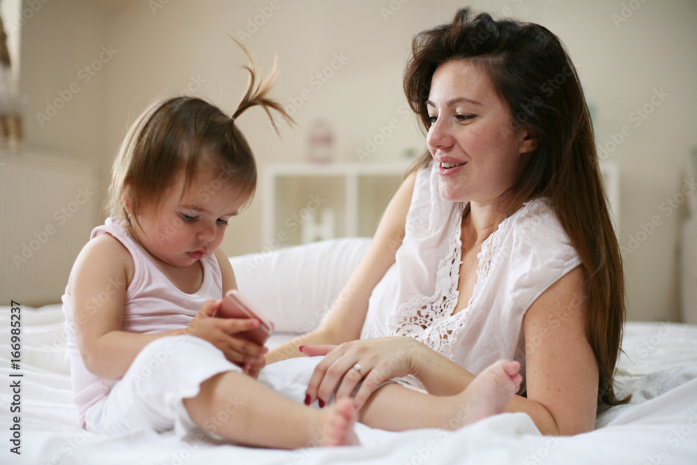 Mother with her little baby having fun in the bed. Using smart phone.