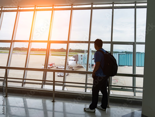 Man looking on the private jet,Asia young man is standing near window at the airport and watching plane before departure. He is standing and carrying luggage. Focus on his back,tourist,trip.