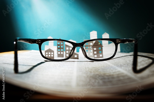 Eyeglasses lie on the open newspaper with paper houses. That could mean rent, search, purchase real estate. 