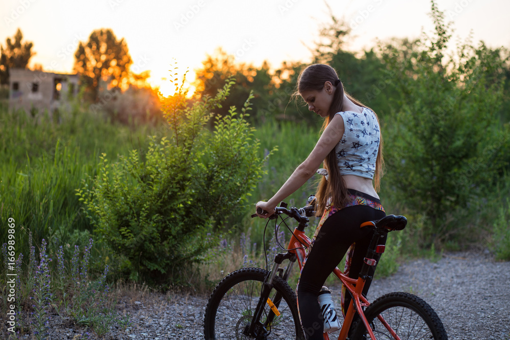 Fototapeta premium Attractive sporty girl with long hair on a bicycle drinking water from the sport bottle on sunset