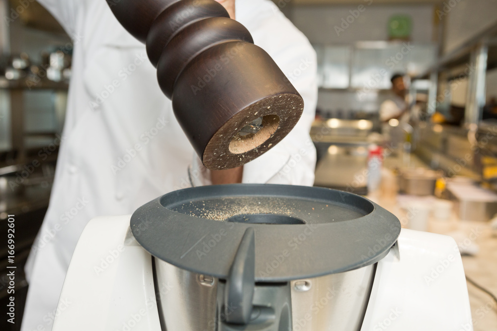 A chef in white chef clothing with a black pepper grinder, grinding fresh  black pepper cloves into a food processor. Stock Photo | Adobe Stock