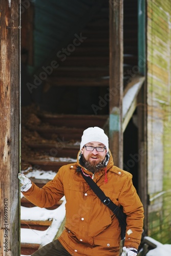 Young smiling red-bearded man stands near the old abandoned houses while traveling in the winter.