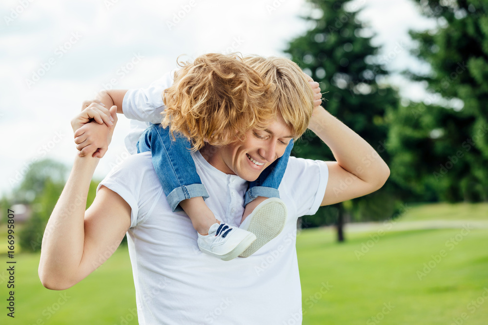 Naklejka premium happy young father carrying adorable little son on neck while walking in park