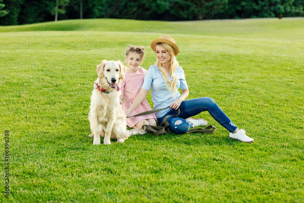 happy beautiful mother and daughter with golden retriever dog sitting on green lawn at park happy beautiful mother and daughter with golden retriever dog sitting on green lawn at park