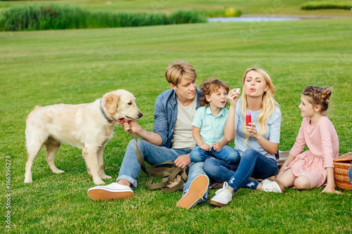 happy young family blowing soap bubbles while resting with dog on grass