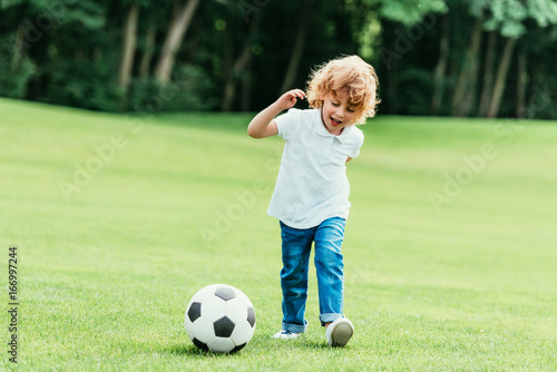 cheerful little boy playing with soccer ball on green lawn at park