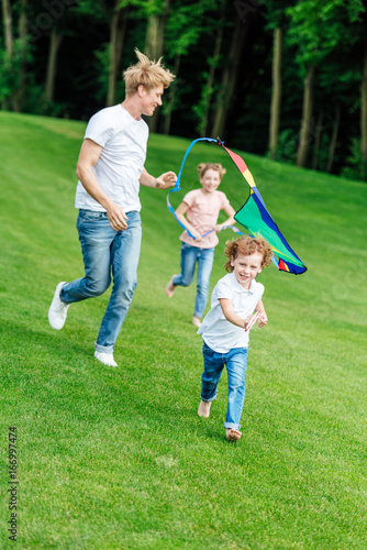 happy father with daughter and son playing with kite while running on green meadow in park