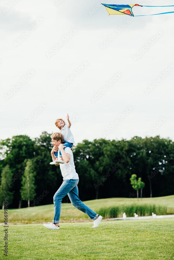 happy father carrying little son on shoulders while playing with kite at park happy father carrying little son on shoulders while playing with kite at park