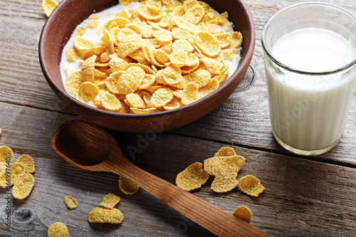 Cornflakes in a brown clay plate on a worn wooden background, next is a glass of milk and a spoon, and several flakes are scattered photo