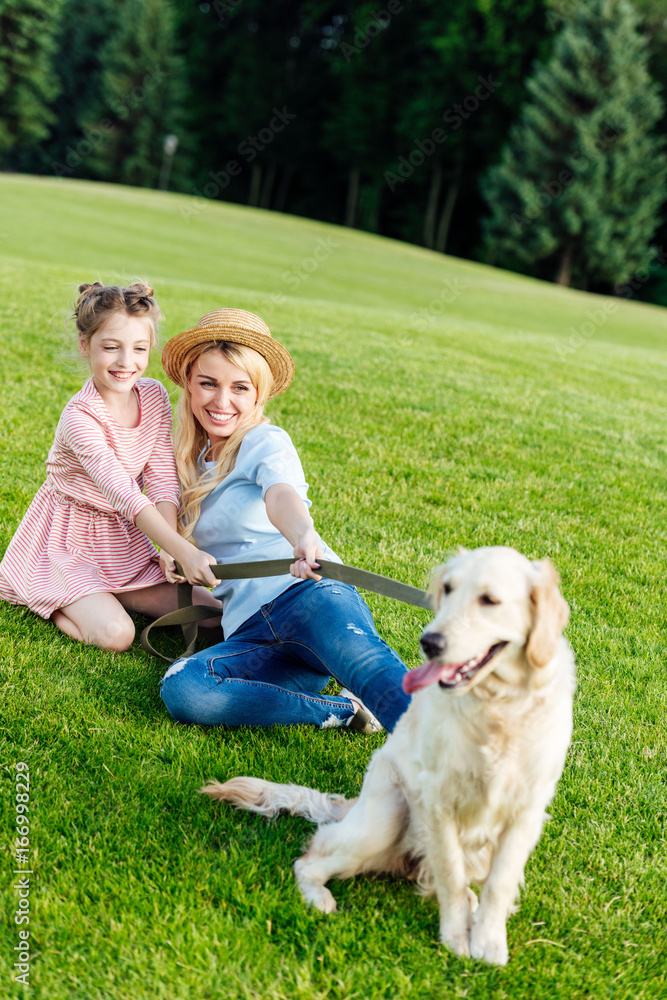 Obraz premium beautiful happy mother and daughter with golden retriever dog resting on green meadow in park