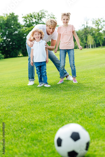 young father with adorable little kids looking at soccer ball on green lawn