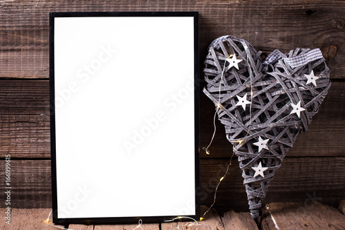 One empty black frame and decorative heart with fairy light on wooden background.