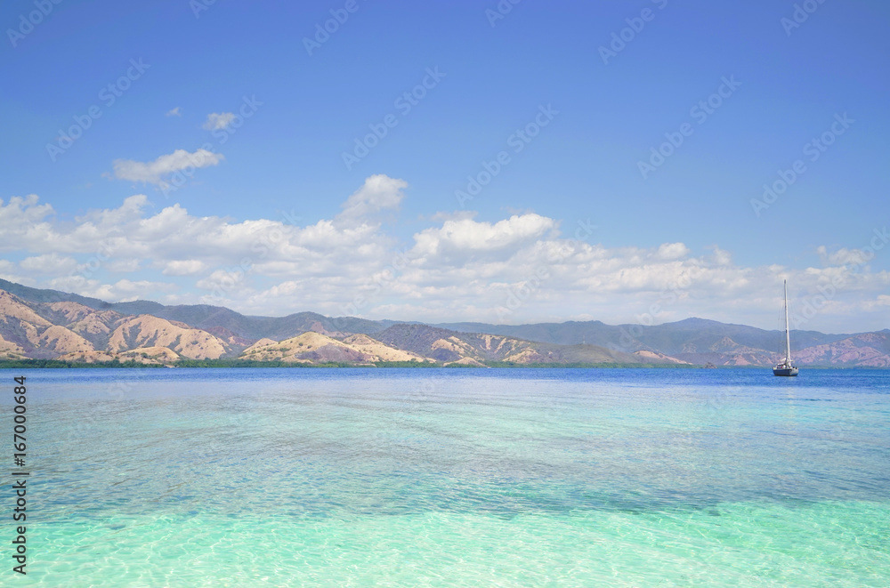 Boat sailing on clear blue tropical ocean water during the day with broad green island behind, Flores Indonesia.
