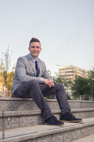one young Caucasian man smiling, business suit, formal wear, ordinary common person portrait, outdoors sitting on stairs steps