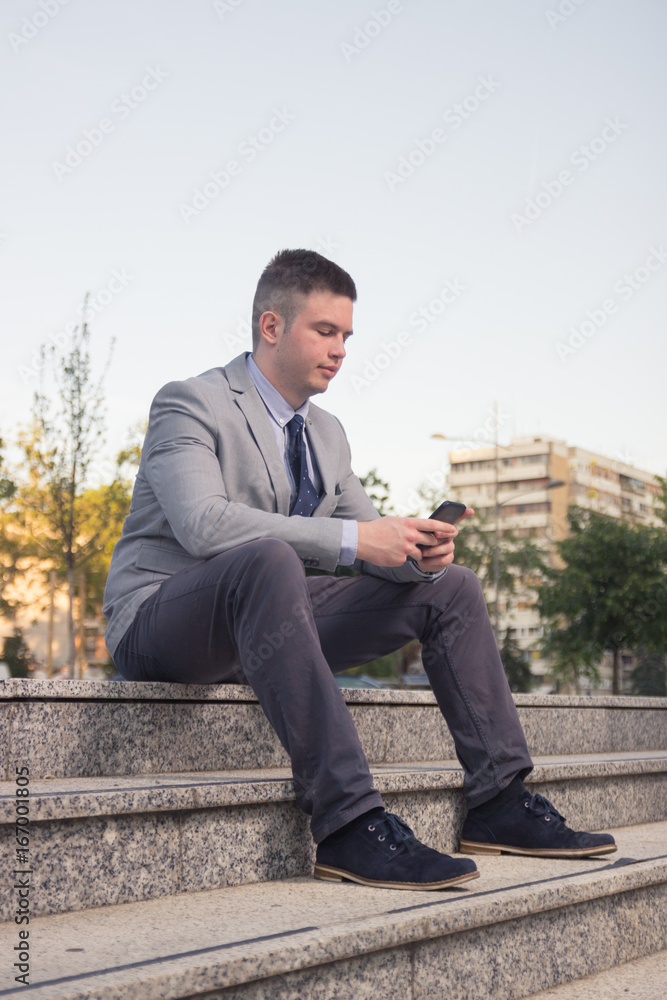 Obraz premium one young Caucasian man smiling, business suit, formal wear, ordinary common person portrait, outdoors sitting on stairs steps, using looking smart phone