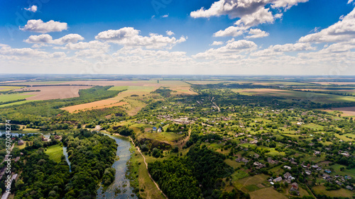 Beautiful view of a typical Ukrainian village surrounded by fields, green forests, small river and blue sky with white clouds. Aerial view.