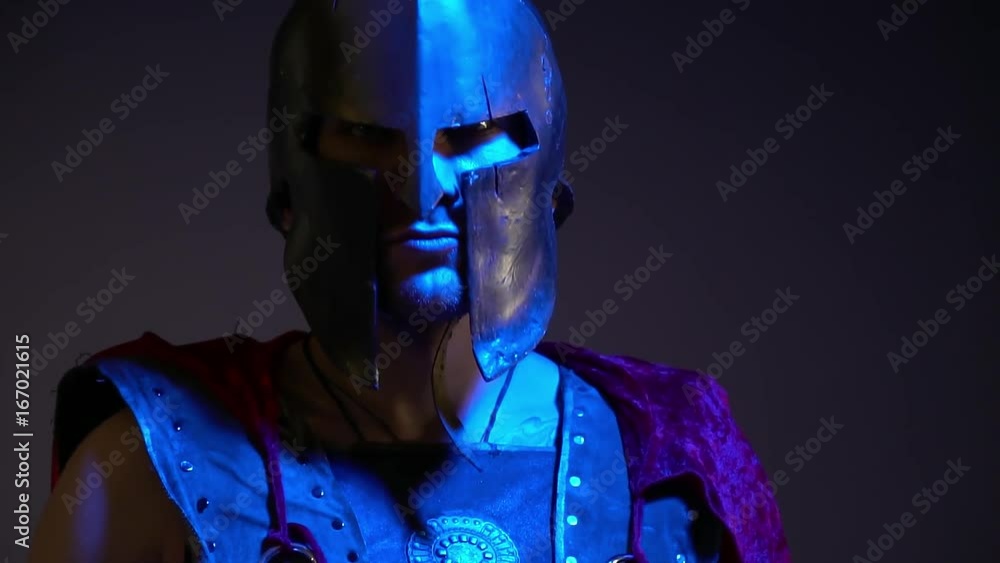 The Roman gladiator in a heavy helmet, leather armor and red raincoat looks into the camera standing in the ultraviolet light, slow motion shooting The Roman gladiator in a heavy helmet, leather armor and red raincoat looks into the camera standing in the ultraviolet light, slow motion shooting