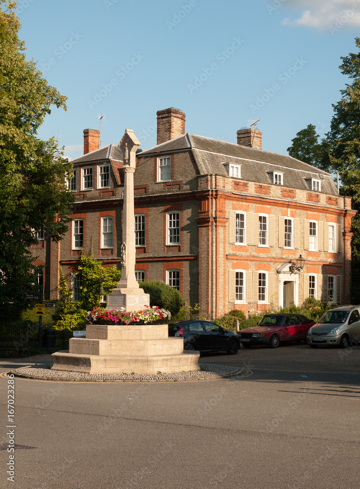 a war memorial landmark in front of a stately home in dedham with some parked cars in summer light