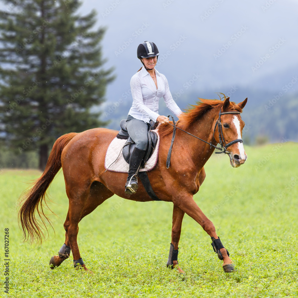 Obraz premium Young woman riding sorrel horse on green mountain meadow. Equestrian activity background