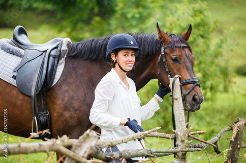 Young smiling rider woman in helmet holding bay horse by bridle