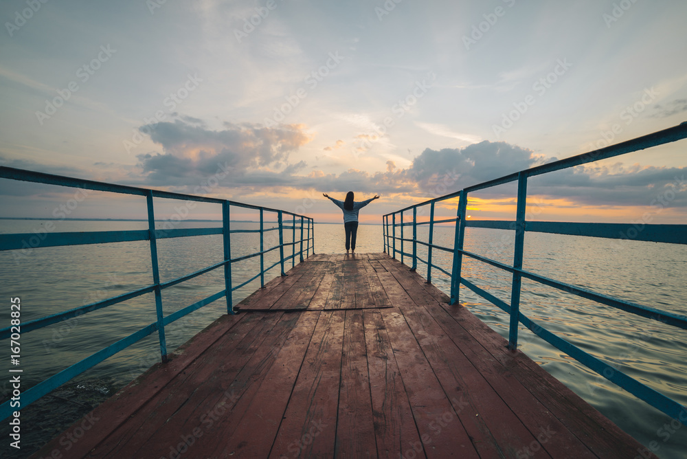 woman stands on the pier on sunrise with hands up
