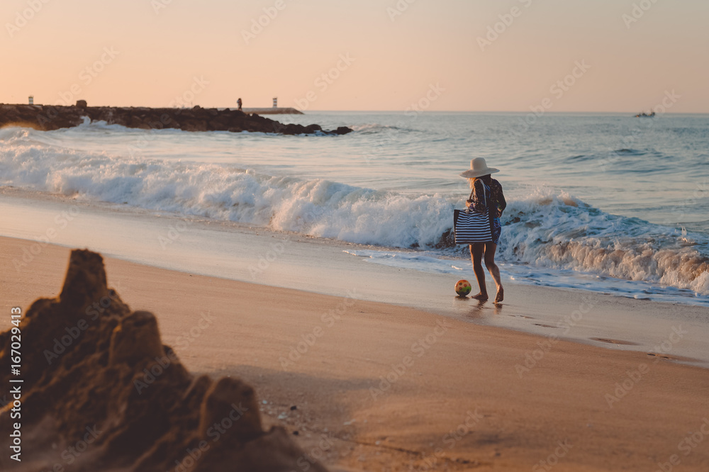 Naklejka premium Holiday lifestyle woman in straw hat on a beach holding bag. Recreational enjoyment walk along sunny outdoors background coastline. Idyllic remote relaxation seashore landscape