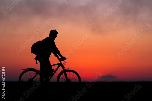 Silhouette of cyclist on the background of red sunset. Biker with bicycle on the field during sunrise