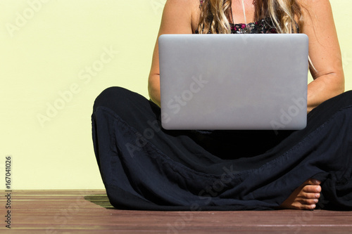  An abstract crop of a beautiful bohemian girl sitting outside on ground with bare feet against a green wall using laptop computer.