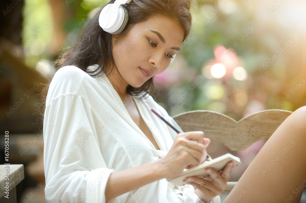 Beautiful young woman take a note,sitting in a garden and headphones on her head Beautiful young woman take a note,sitting in a garden and headphones on her head