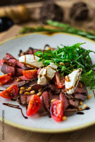 On a plate, on a wooden surface, close-up roast beef salad with rocket salad, cherry tomatoes, mozzarella, balsamic sauce, rosemary, spices, nuts, tall kitchen, restaurant, trendy delicious food