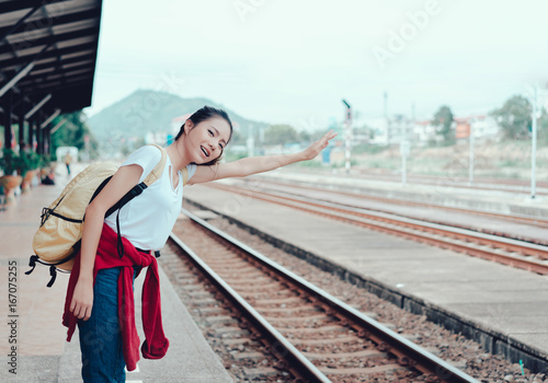 Young girl asian tourist standing with backpack waiting the train going to travel scenery town tour around at train station platform for take the rest, happy and life experience having fun