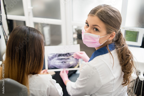 Female young dentist examining x-ray image with female patient in dental office  looking to the camera and preparing for treatment. Dentistry