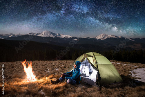 Female tourist have a rest in her camp at night. Happy woman sitting near campfire and a green tent under beautiful sky full of stars and milky way. On the background snow-covered mountains