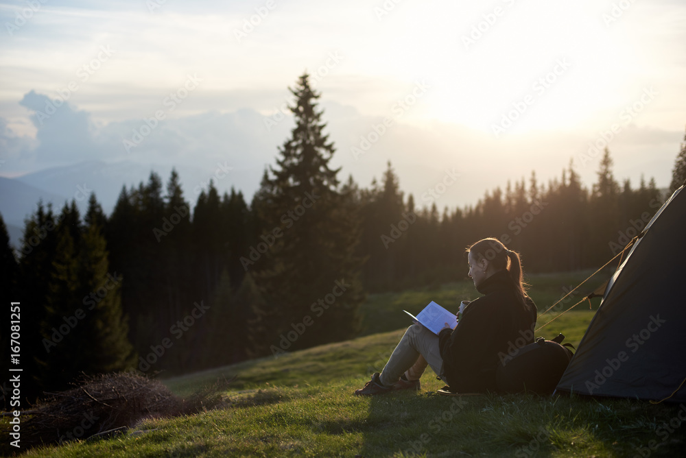 Young woman tourist sitting near the tent and reading a book at sunset in the mountains