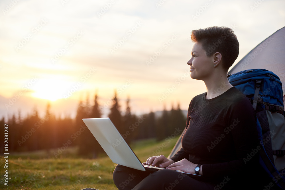 Happy woman looking away thoughtfully using her laptop outdoors while camping sitting near her tent dreaming achievement success travel online connection