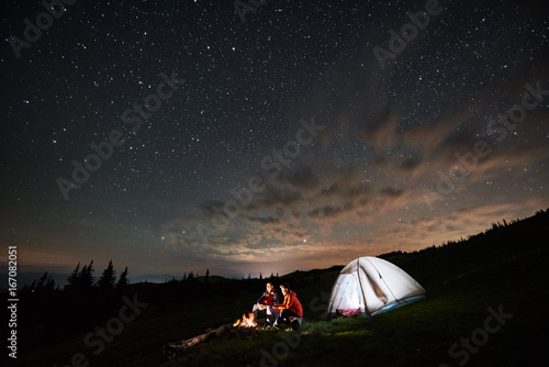 Night camping in the mountains. Couple tourists have a rest at a campfire near illuminated tent under amazing night starry sky. Low light