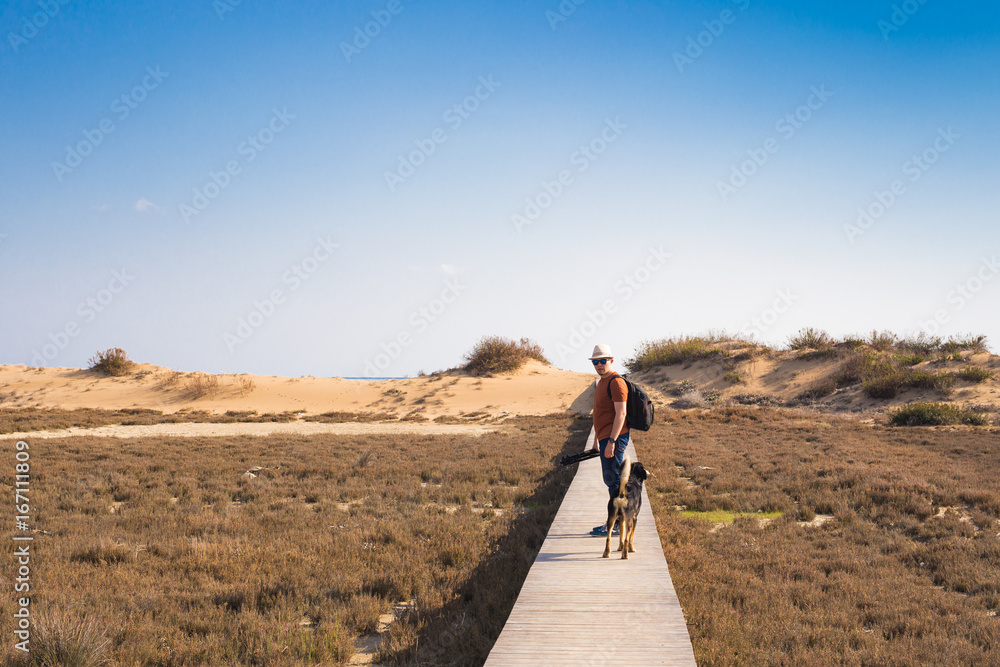 Man with dog walking on the wooden path on the beach and looking into the distance of the ocean