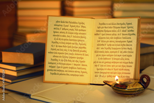 Open book illuminated by an oil lamp. The opening lines of The Iliad written in the ancient Greek language. A stack of books. On the lamp, visible inscription Aphrodite in the Greek alphabet. photo