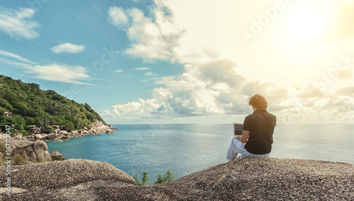 Young man, the freelancer sits on a high cliff over the sea and works remotely