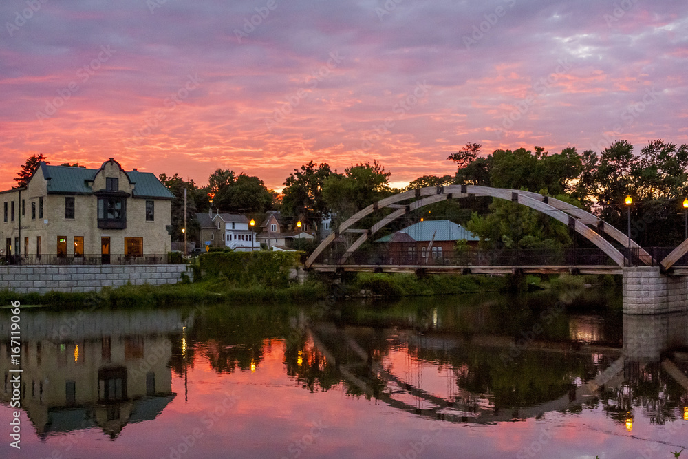 Fototapeta premium A very colorful sunset over Jefferson, Wisconsin reflected in the Rock River with the arched walk bridge.