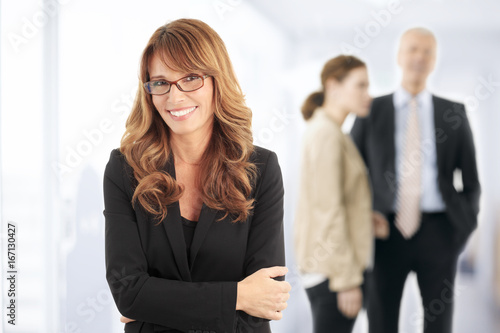 Shot of a beautiful middle aged financial businesswoman standing with arms crossed in the office while business people consulting in the background.