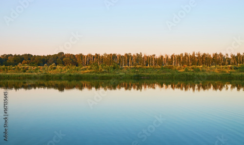 The blue sky and trees are reflected on the surface of the river at sunset.