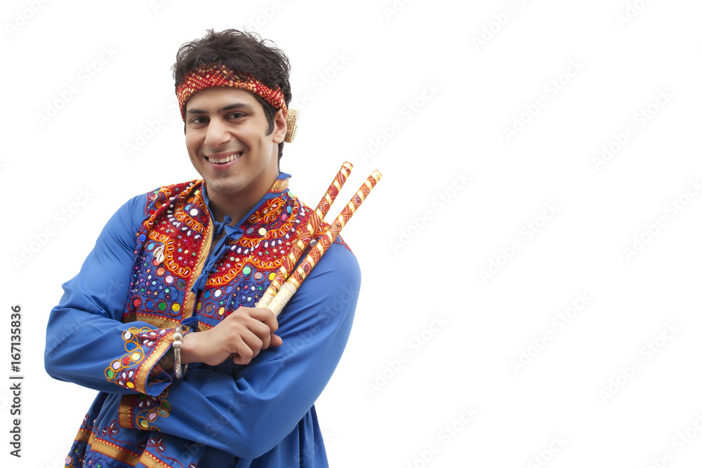 Portrait of happy young man in traditional wear holding dandiya over white background Portrait of happy young man in traditional wear holding dandiya over white background