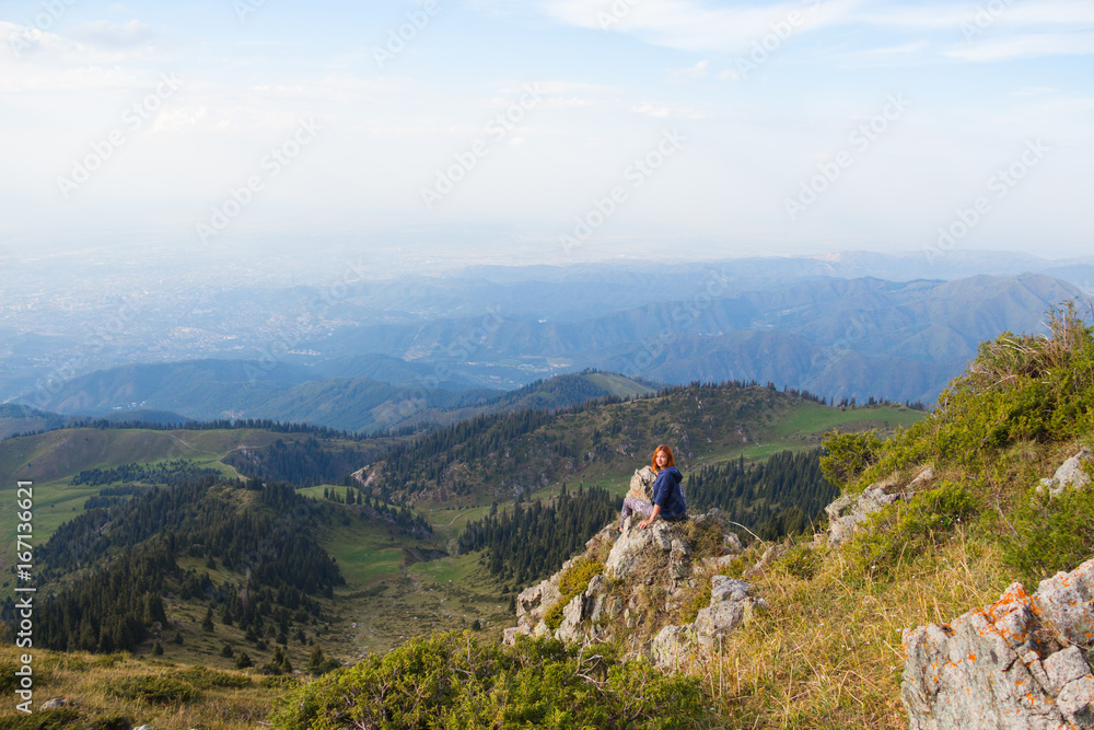 Mountain view with young woman sitting on top. Tourism concept background.