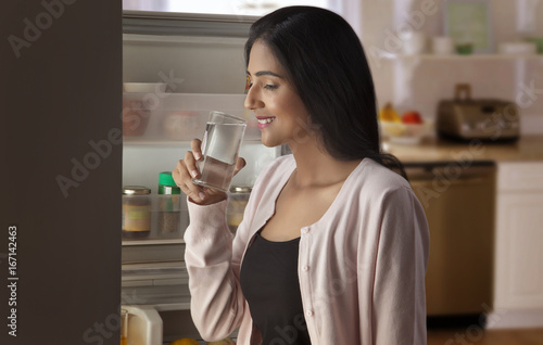 Young woman drinking water in front of open refrigerator 